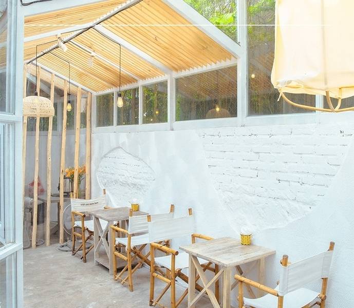 Qu&aacute;n c&agrave; ph&ecirc; với b&agrave;n ghế gỗ, tường trắng v&agrave; m&aacute;i k&iacute;nh, &aacute;nh s&aacute;ng tự nhi&ecirc;n từ cửa sổ lớn.