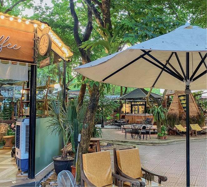 Qu&aacute;n c&agrave; ph&ecirc; ngo&agrave;i trời với c&acirc;y xanh, b&agrave;n ghế, d&ugrave; che v&agrave; kh&ocirc;ng gian tho&aacute;ng đ&atilde;ng, trang tr&iacute; ấm c&uacute;ng.