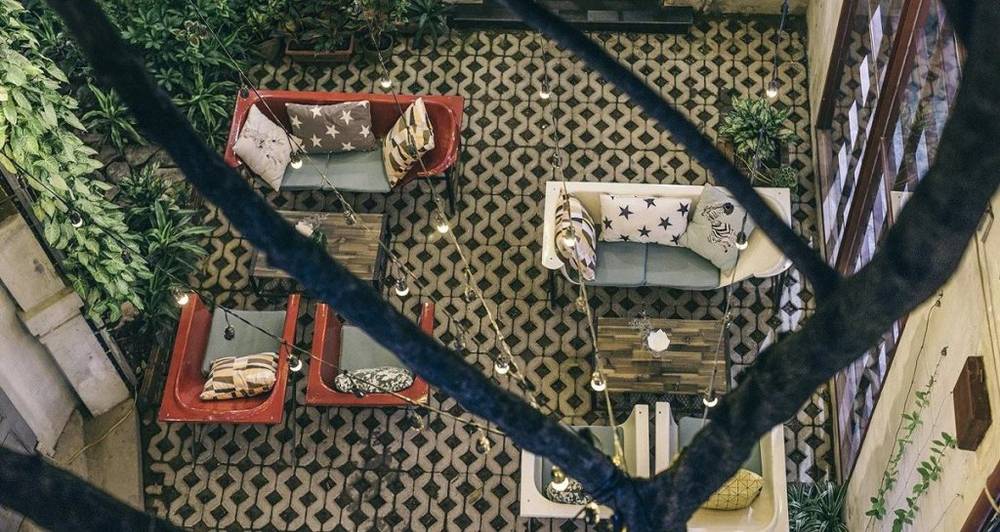 Khu vực ngồi ngo&agrave;i trời với b&agrave;n ghế v&agrave; đệm trang tr&iacute;, l&aacute;t gạch hoa văn, c&oacute; c&acirc;y xanh xung quanh.