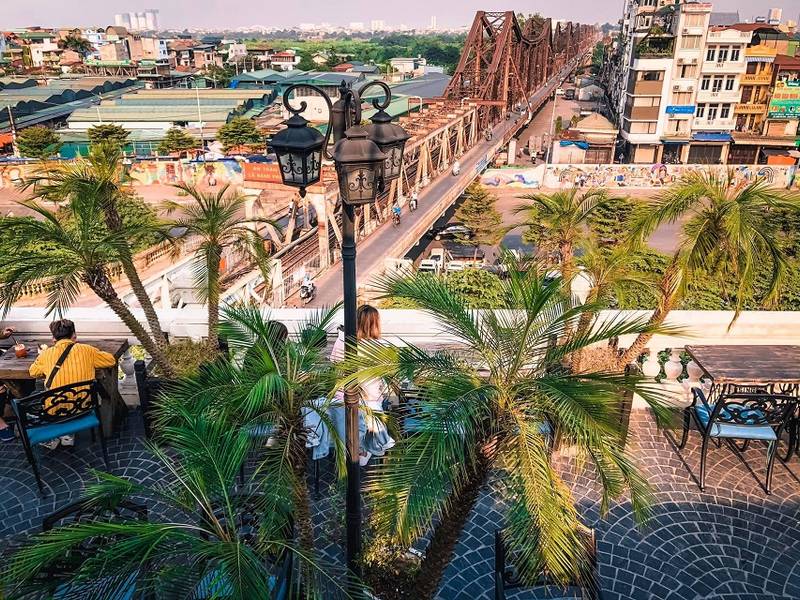 Quang cảnh một qu&aacute;n c&agrave; ph&ecirc; tr&ecirc;n cao với tầm nh&igrave;n ra c&acirc;y cầu sắt v&agrave; c&aacute;c t&ograve;a nh&agrave; xung quanh, c&oacute; c&acirc;y cọ v&agrave; đ&egrave;n trang tr&iacute;.