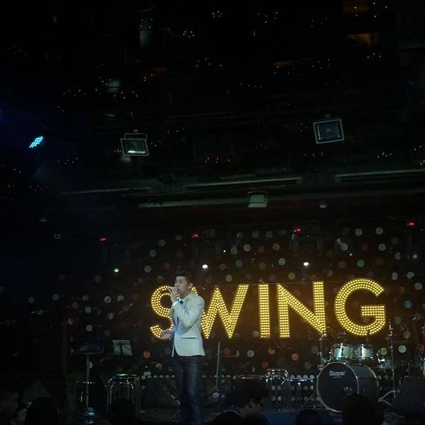 Người đang biểu diễn tr&ecirc;n s&acirc;n khấu với &aacute;nh đ&egrave;n chữ "SWING" ph&iacute;a sau, c&oacute; nhạc cụ b&ecirc;n cạnh.