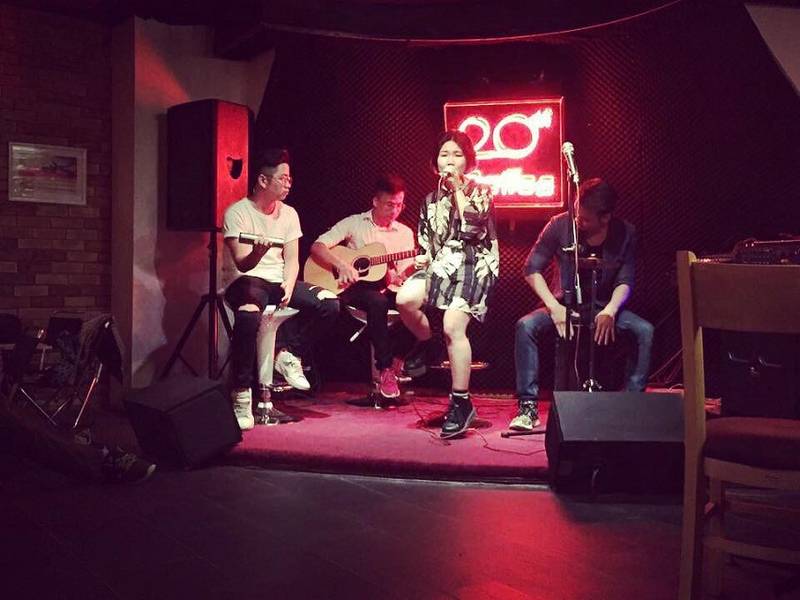 Ban nhạc biểu diễn tr&ecirc;n s&acirc;n khấu nhỏ với &aacute;nh s&aacute;ng mờ, c&oacute; người chơi guitar v&agrave; h&aacute;t trước biển hiệu neon.
