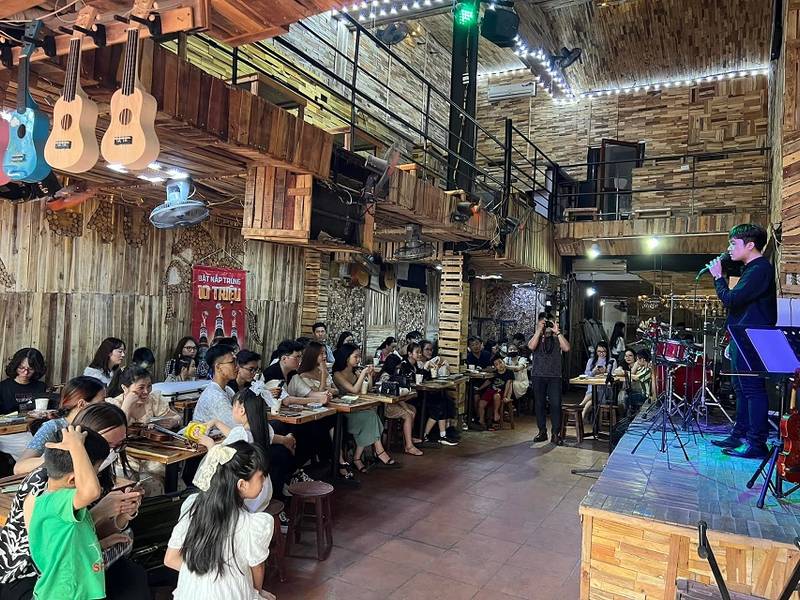 Qu&aacute;n c&agrave; ph&ecirc; với s&acirc;n khấu biểu diễn, đ&ocirc;ng người ngồi xem nghệ sĩ h&aacute;t tr&ecirc;n s&acirc;n khấu gỗ, trang tr&iacute; nhạc cụ tr&ecirc;n tường.