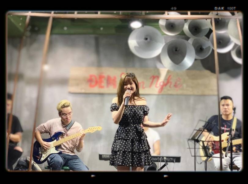 Ca sĩ biểu diễn tr&ecirc;n s&acirc;n khấu với ban nhạc, người chơi guitar v&agrave; bass, nền c&oacute; d&ograve;ng chữ "Đ&ecirc;m Văn Nghệ".