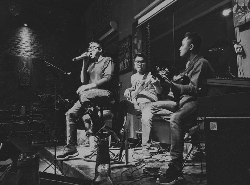 Nh&oacute;m nhạc biểu diễn tr&ecirc;n s&acirc;n khấu nhỏ, một người h&aacute;t, hai người chơi guitar trong kh&ocirc;ng gian qu&aacute;n bar tối.