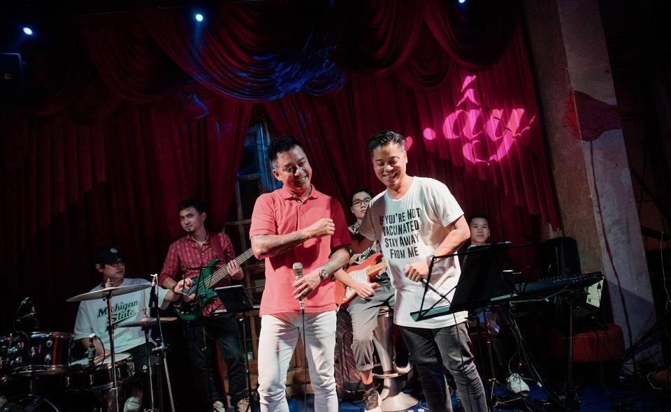 Nh&oacute;m nhạc biểu diễn tr&ecirc;n s&acirc;n khấu với một người cầm micro, ph&iacute;a sau l&agrave; c&aacute;c nhạc cụ v&agrave; ph&ocirc;ng nền đỏ.