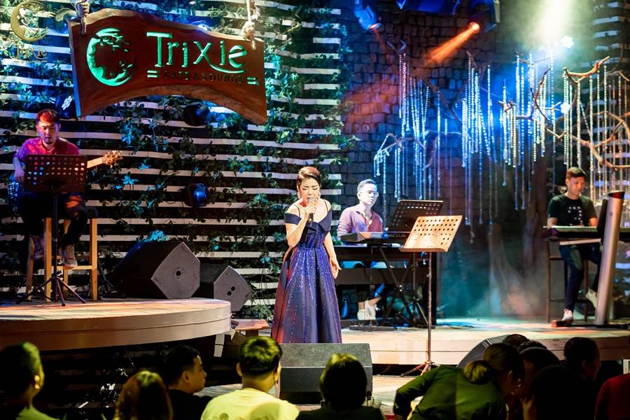 Ca sĩ biểu diễn tr&ecirc;n s&acirc;n khấu c&ugrave;ng ban nhạc trong kh&ocirc;ng gian qu&aacute;n c&agrave; ph&ecirc;, &aacute;nh đ&egrave;n nhiều m&agrave;u sắc.