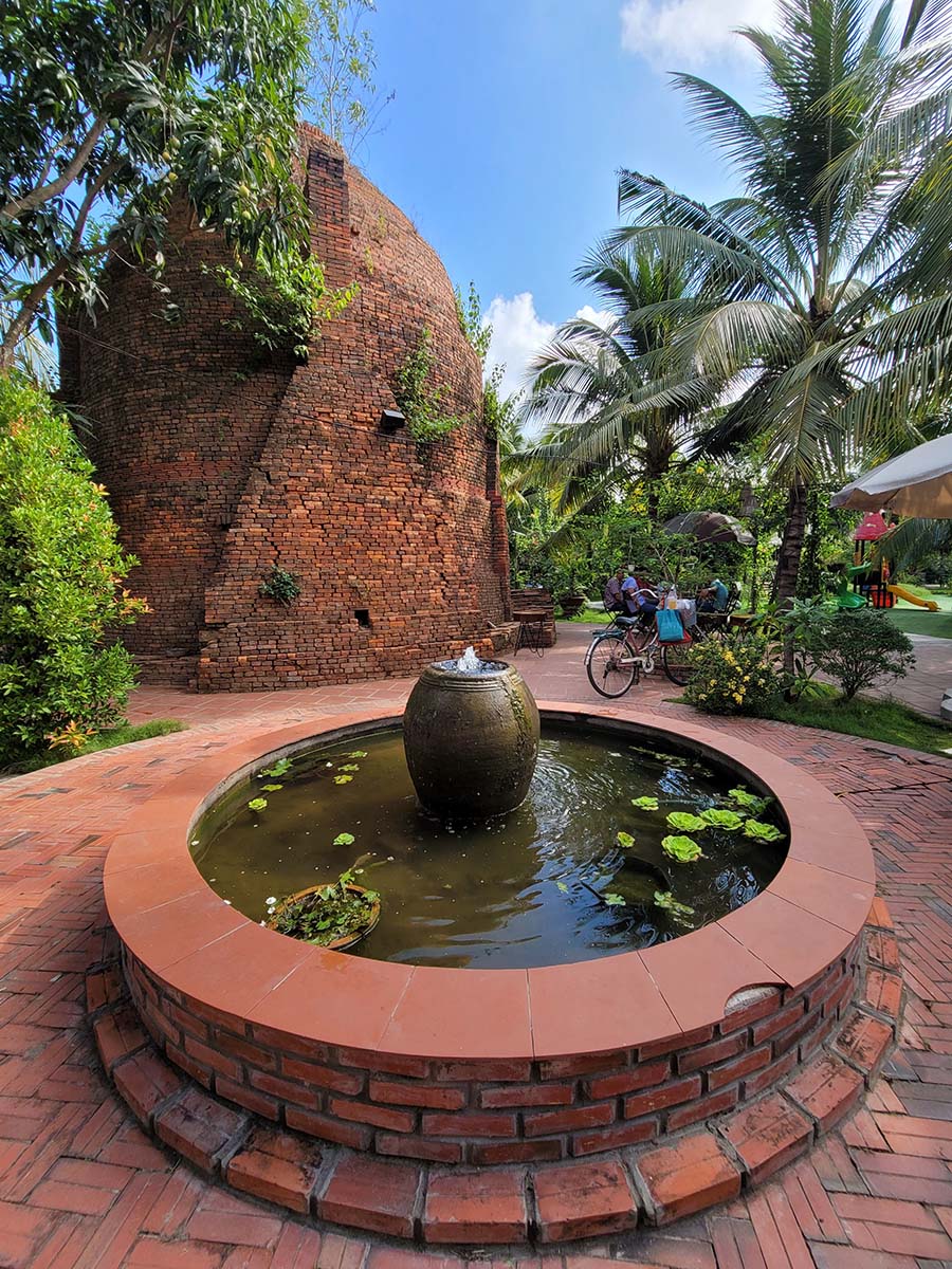 Khu vườn c&oacute; bức tường gạch cổ v&agrave; đ&agrave;i phun nước nhỏ, bao quanh bởi c&acirc;y cối xanh tươi v&agrave; bầu trời trong xanh.