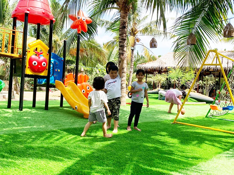 Trẻ em vui chơi tr&ecirc;n s&acirc;n chơi ngo&agrave;i trời với cầu trượt v&agrave; x&iacute;ch đu, xung quanh l&agrave; c&acirc;y cọ v&agrave; cỏ xanh.