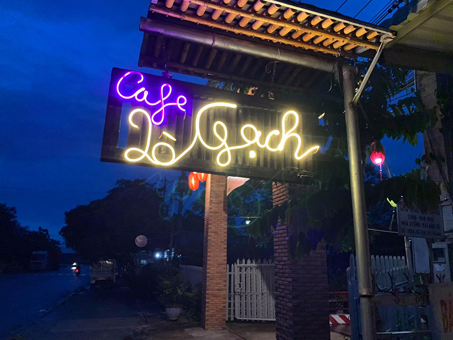 Biển hiệu qu&aacute;n c&agrave; ph&ecirc; với chữ neon m&agrave;u t&iacute;m v&agrave; v&agrave;ng, treo dưới m&aacute;i hi&ecirc;n v&agrave;o buổi tối.