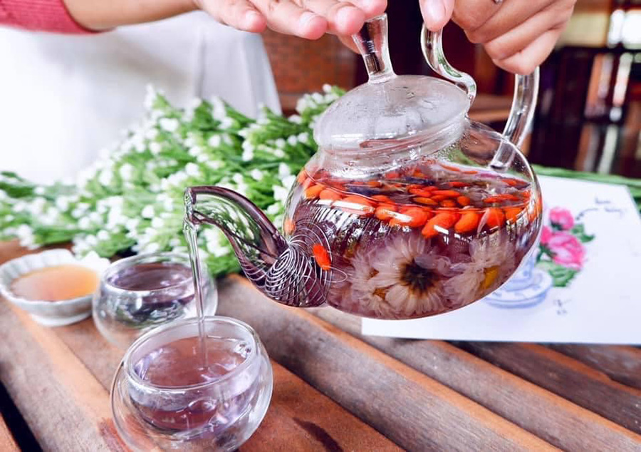 Ấm tr&agrave; thủy tinh với hoa v&agrave; quả, đang r&oacute;t v&agrave;o ly, b&ecirc;n cạnh l&agrave; hoa trắng tr&ecirc;n b&agrave;n gỗ.