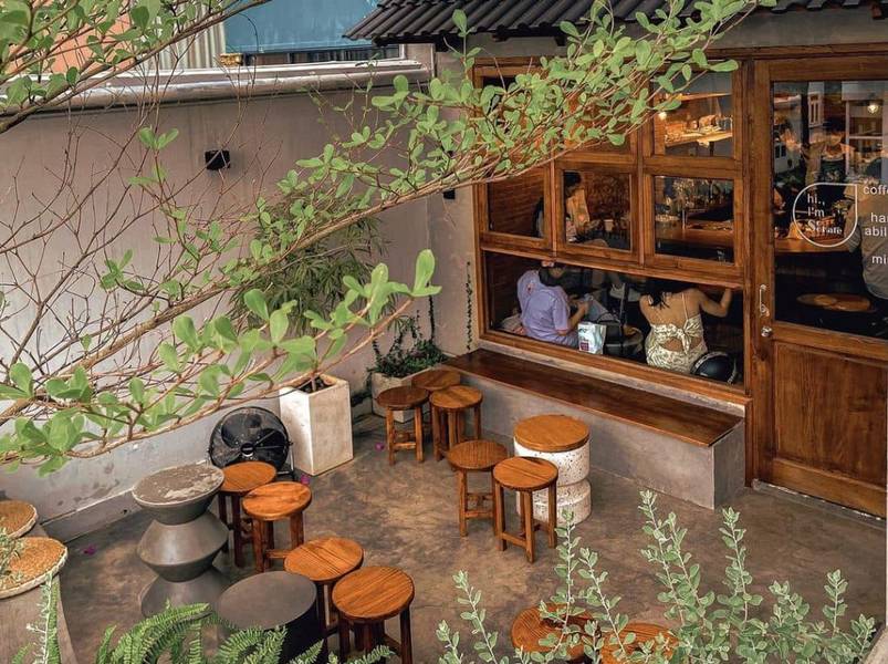 Quán cà phê ngoài trời với bàn ghế gỗ, cây xanh bao quanh, cửa kính lớn nhìn vào bên trong.