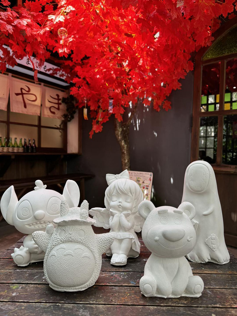 C&aacute;c bức tượng thạch cao h&igrave;nh th&uacute; v&agrave; nh&acirc;n vật hoạt h&igrave;nh tr&ecirc;n b&agrave;n gỗ, dưới t&aacute;n c&acirc;y l&aacute; đỏ.