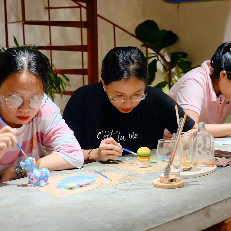 Ba người đang ngồi vẽ m&ocirc; h&igrave;nh tại một b&agrave;n, xung quanh c&oacute; cọ vẽ v&agrave; ly nước.