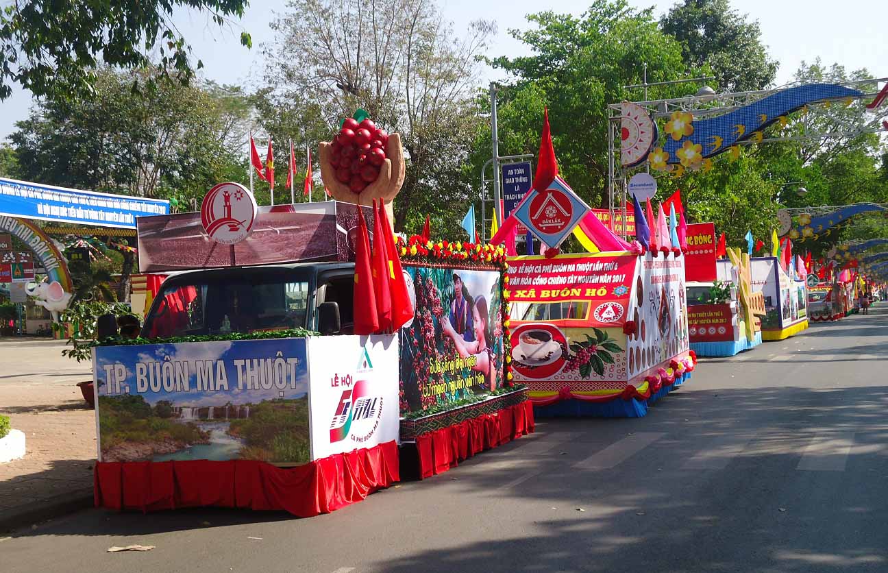 Xe diễu hành trang trí màu sắc và biểu ngữ, tham gia lễ hội trên đường phố, có nhiều cây và cờ xung quanh.