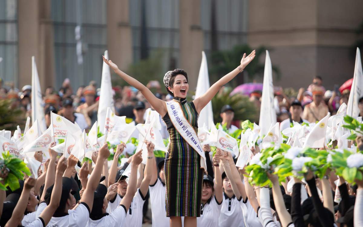 Người phụ nữ đứng giữa đám đông đang giơ cao cờ, mặc trang phục truyền thống và đeo dải băng chéo.