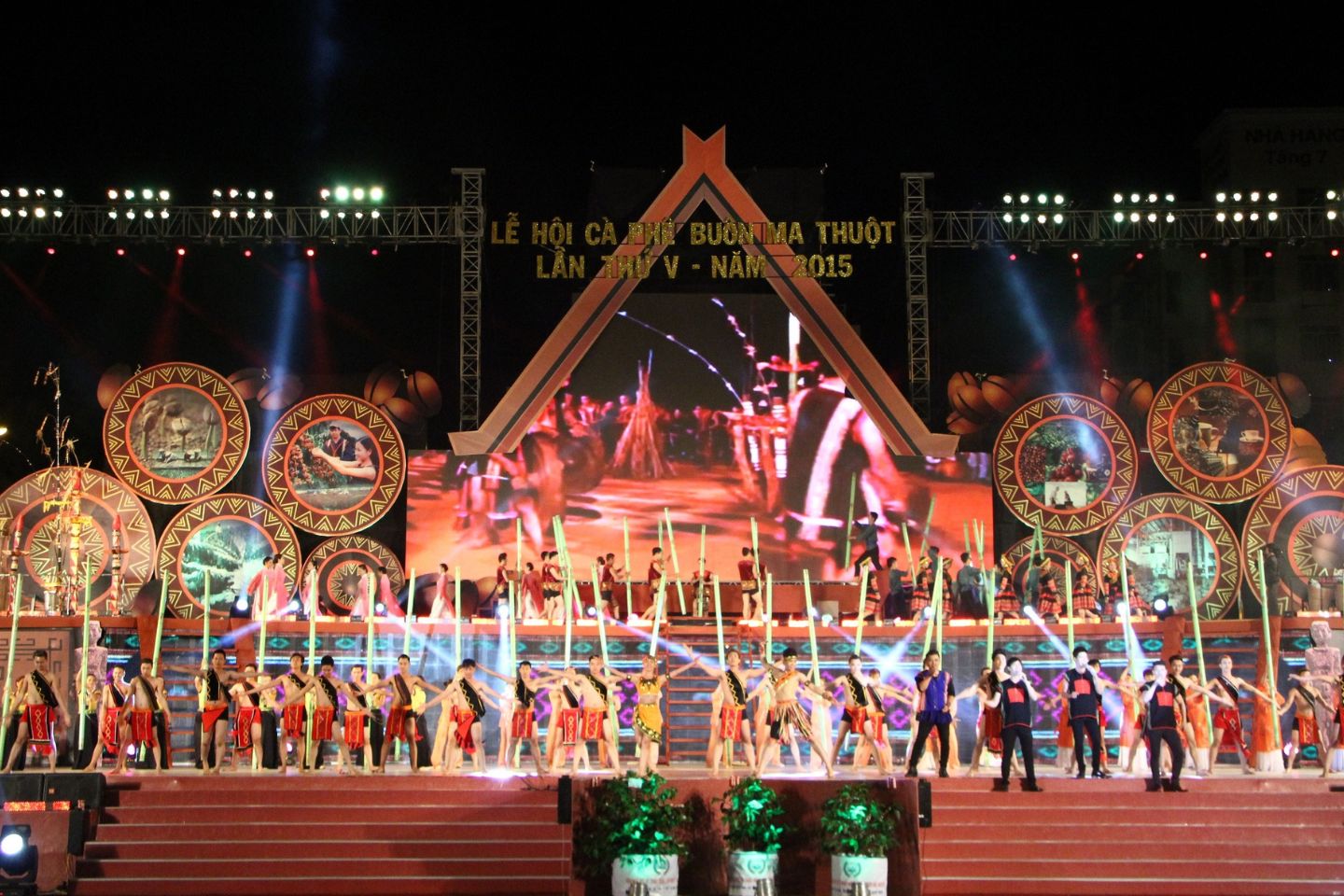 Sân khấu trình diễn với nhiều người trong trang phục truyền thống, nền là màn hình lớn và ánh sáng rực rỡ.