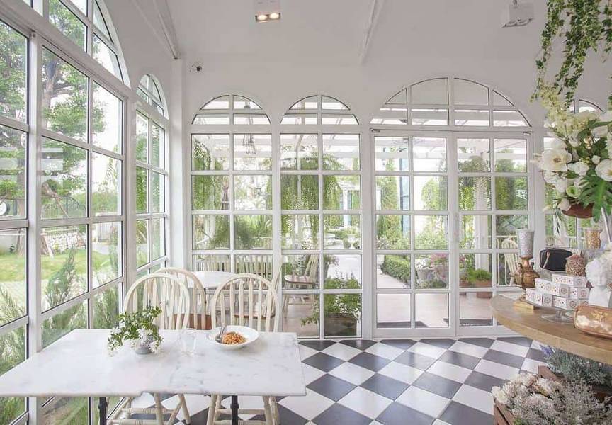 Qu&aacute;n c&agrave; ph&ecirc; s&aacute;ng sủa với cửa k&iacute;nh lớn, b&agrave;n ghế gỗ trắng v&agrave; c&acirc;y xanh trang tr&iacute; xung quanh.