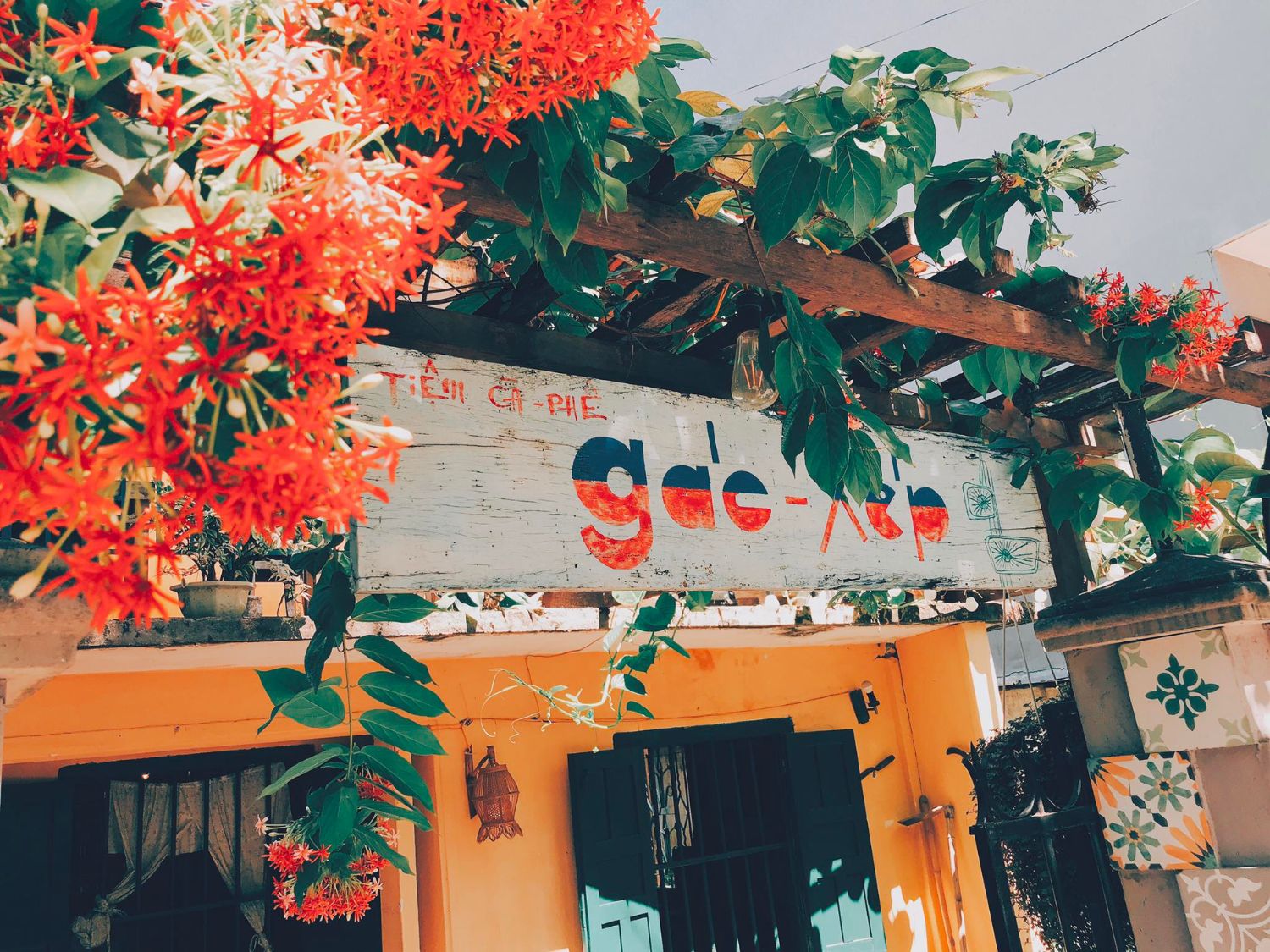 Qu&aacute;n c&agrave; ph&ecirc; nhỏ với bảng hiệu gỗ, xung quanh trang tr&iacute; hoa giấy đỏ rực v&agrave; c&acirc;y xanh.