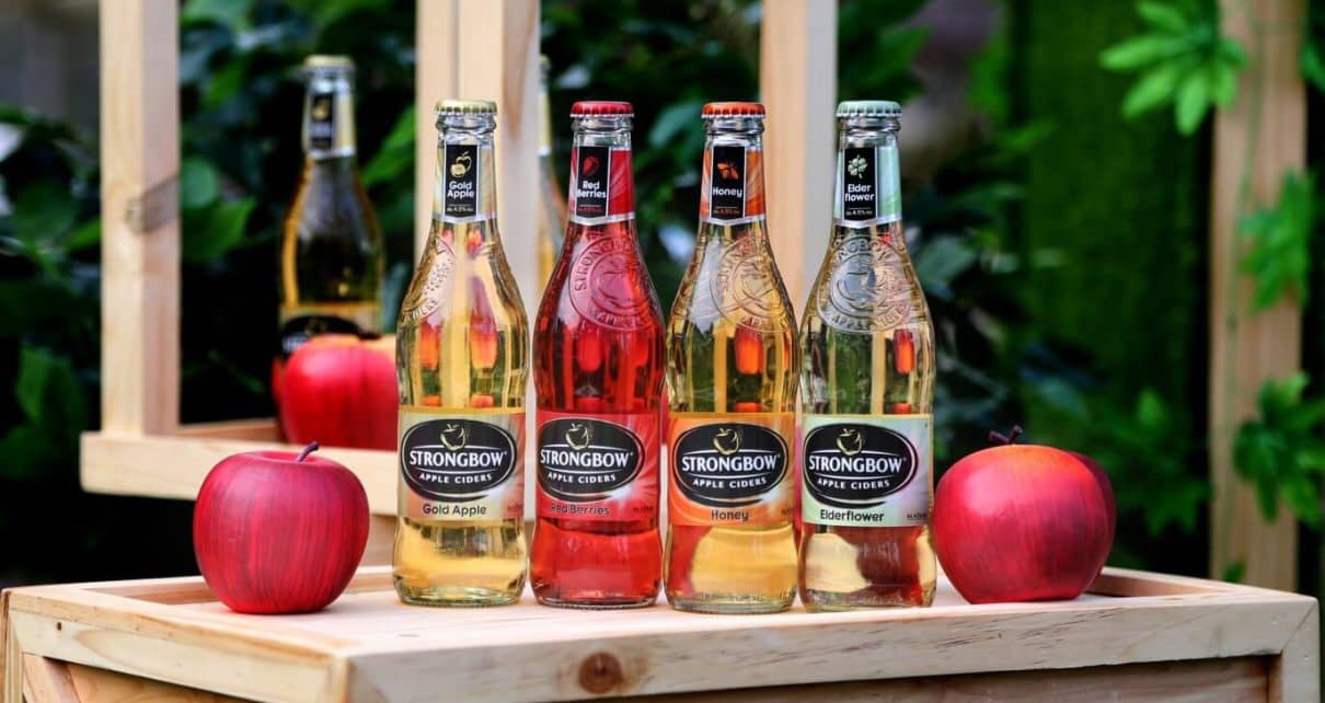 Bốn chai cider Strongbow với c&aacute;c hương vị kh&aacute;c nhau được đặt tr&ecirc;n hộp gỗ, b&ecirc;n cạnh l&agrave; hai quả t&aacute;o trang tr&iacute;.