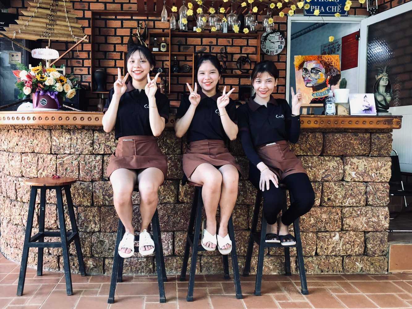 Ba người ngồi tr&ecirc;n ghế cao trước quầy bar gạch, mặc đồng phục, cười v&agrave; giơ tay tạo d&aacute;ng chữ V.