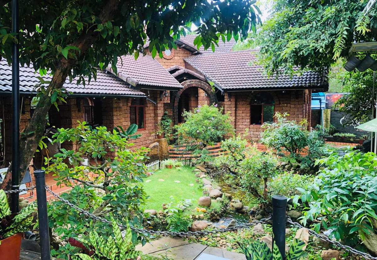 Ng&ocirc;i nh&agrave; gạch đỏ giữa vườn c&acirc;y xanh tươi, c&oacute; m&aacute;i ng&oacute;i v&agrave; lối đi nhỏ, tạo cảnh quan y&ecirc;n b&igrave;nh v&agrave; gần gũi thi&ecirc;n nhi&ecirc;n.