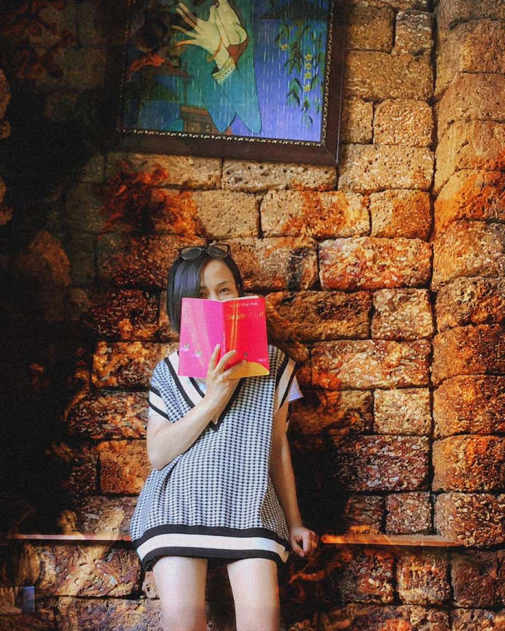 Người phụ nữ ngồi dựa v&agrave;o tường gạch, che mặt bằng cuốn s&aacute;ch hồng, tr&ecirc;n đầu l&agrave; bức tranh nghệ thuật.