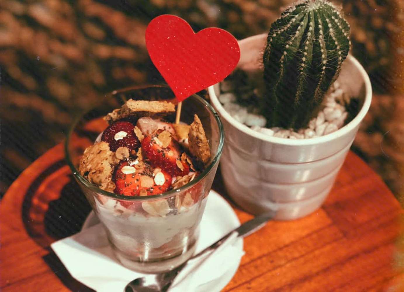 Ly sữa chua tr&aacute;i c&acirc;y trang tr&iacute; tim đỏ, cạnh c&acirc;y xương rồng trong chậu tr&ecirc;n b&agrave;n gỗ.