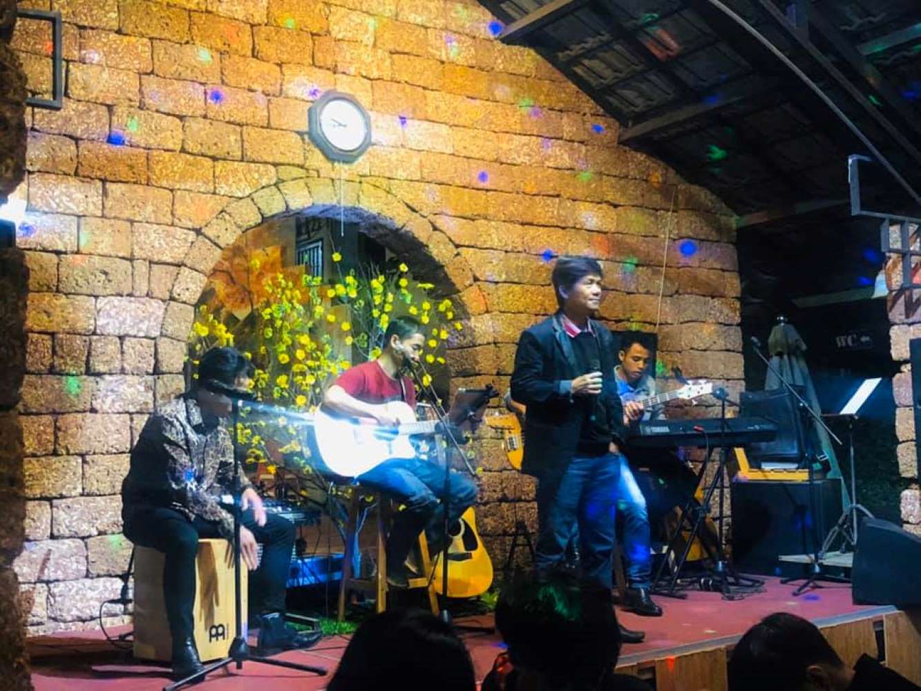 Ban nhạc biểu diễn tr&ecirc;n s&acirc;n khấu với tường gạch v&agrave; hoa v&agrave;ng ph&iacute;a sau, c&oacute; người h&aacute;t v&agrave; chơi nhạc cụ.