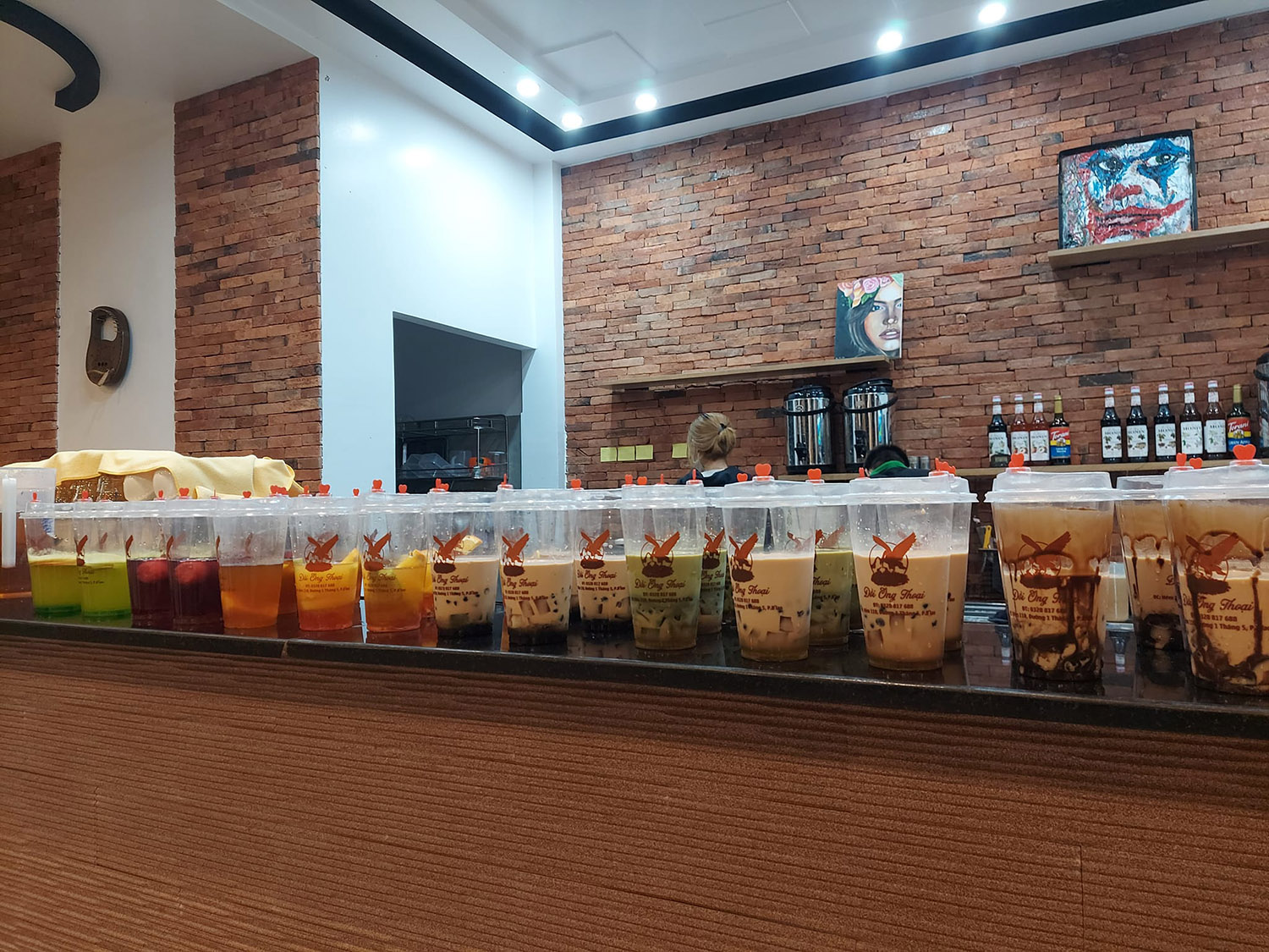 H&agrave;ng loạt ly tr&agrave; sữa v&agrave; nước uống với nhiều m&agrave;u sắc tr&ecirc;n quầy, nền tường gạch, trang tr&iacute; tranh nghệ thuật.