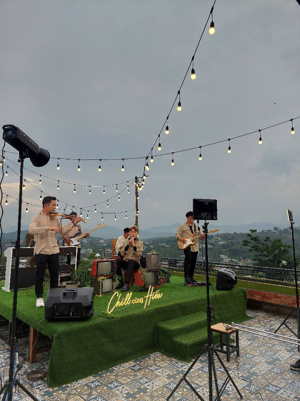 Ban nhạc biểu diễn tr&ecirc;n s&acirc;n khấu ngo&agrave;i trời với &aacute;nh đ&egrave;n treo, nền trời xanh v&agrave; cảnh quan thi&ecirc;n nhi&ecirc;n ph&iacute;a xa.