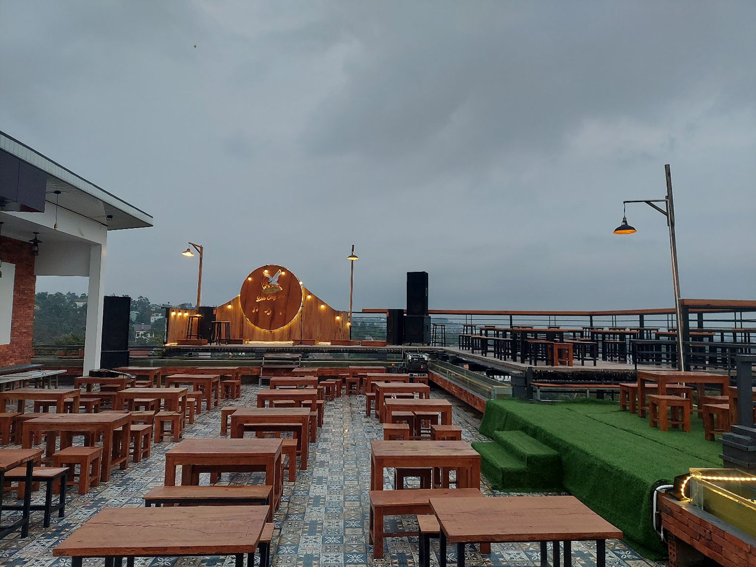 Qu&aacute;n c&agrave; ph&ecirc; s&acirc;n thượng với b&agrave;n ghế gỗ, s&acirc;n khấu nhỏ c&oacute; đ&egrave;n trang tr&iacute;, bầu trời &acirc;m u ph&iacute;a tr&ecirc;n.