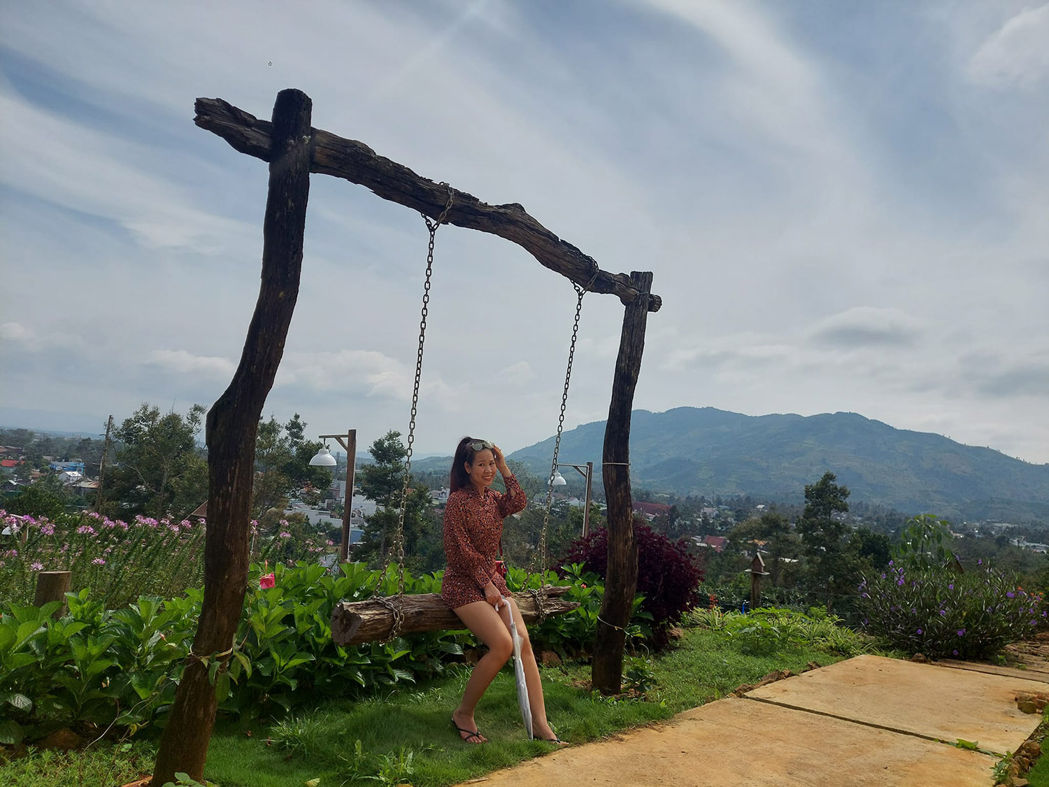 Người ngồi tr&ecirc;n x&iacute;ch đu gỗ giữa vườn hoa, nền l&agrave; n&uacute;i non v&agrave; bầu trời xanh.