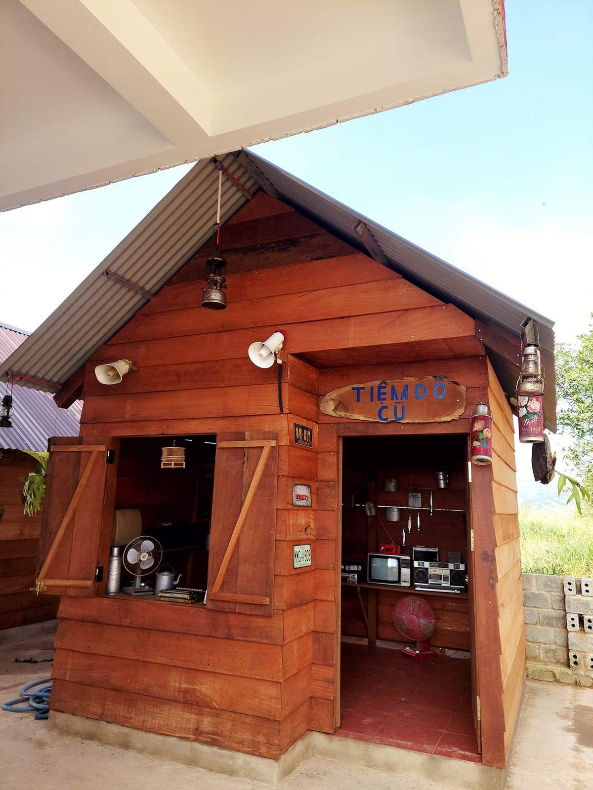 Gian nh&agrave; gỗ nhỏ với biển hiệu ph&iacute;a tr&ecirc;n cửa, b&ecirc;n trong c&oacute; quạt, tivi v&agrave; c&aacute;c vật dụng điện tử cũ.