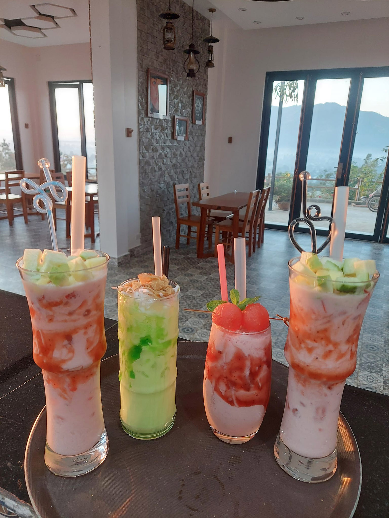 Bốn ly đồ uống tr&aacute;i c&acirc;y trang tr&iacute; đẹp mắt tr&ecirc;n khay, đặt trong kh&ocirc;ng gian qu&aacute;n c&oacute; cửa sổ lớn nh&igrave;n ra ngo&agrave;i.