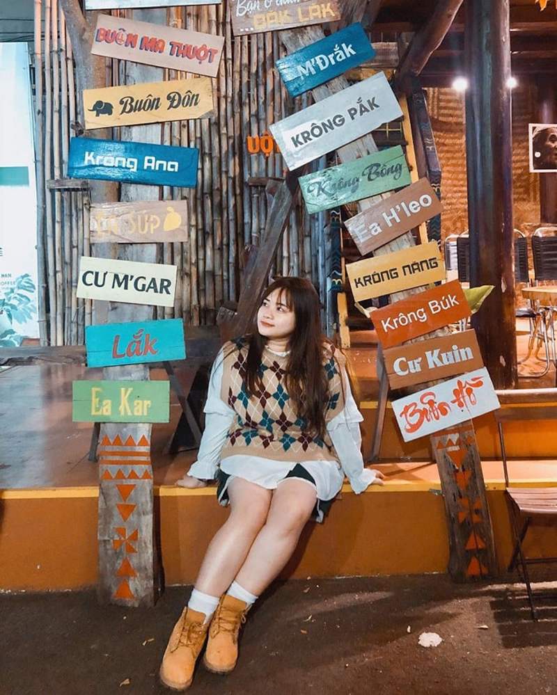 Người ngồi cạnh bảng chỉ đường với nhiều tên địa điểm, nền gỗ và ánh sáng ấm áp.