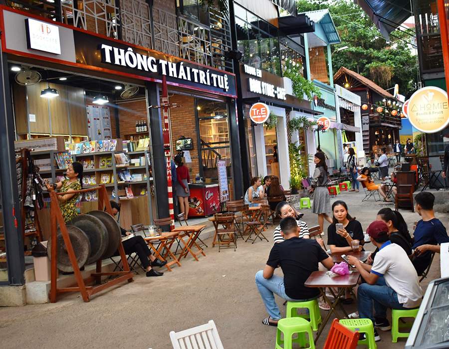 Khu vực quán cà phê ngoài trời với nhiều người ngồi trò chuyện bên các bàn ghế nhỏ trước cửa hàng sách.