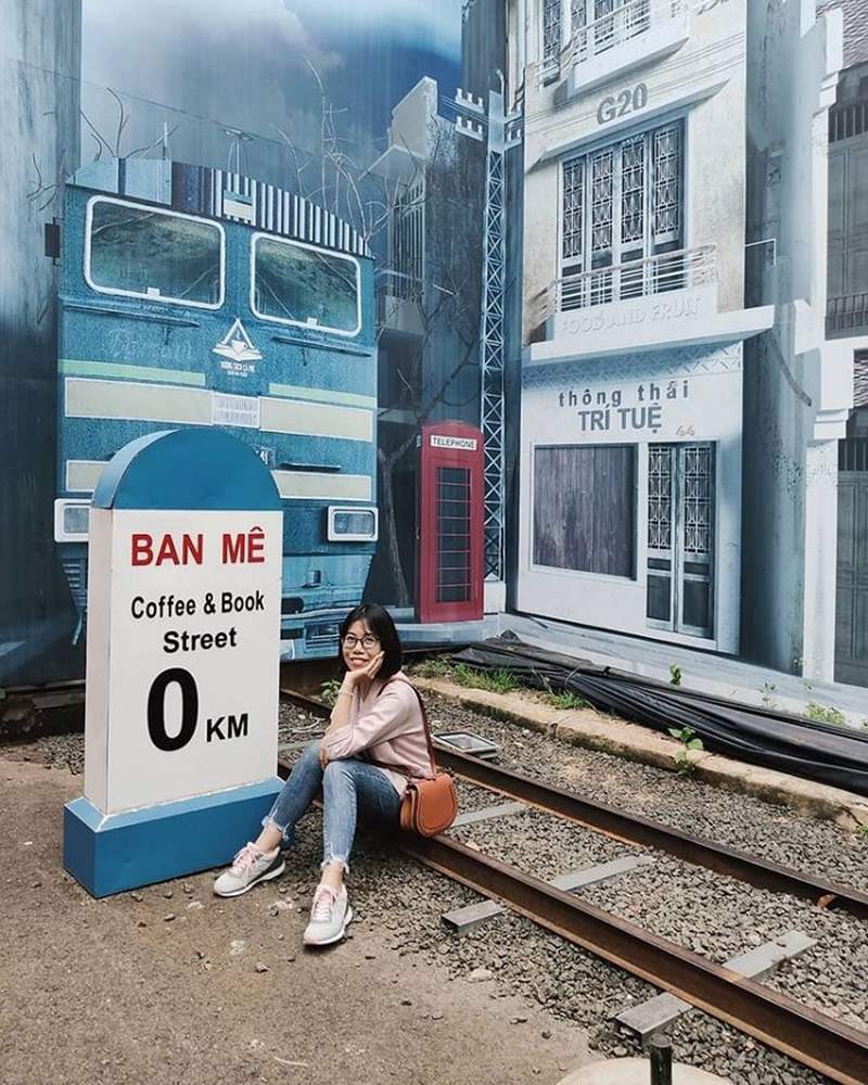 Người ngồi trên đường ray trước bức tranh xe lửa và biển báo "Ban Mê Coffee & Book Street 0 KM".