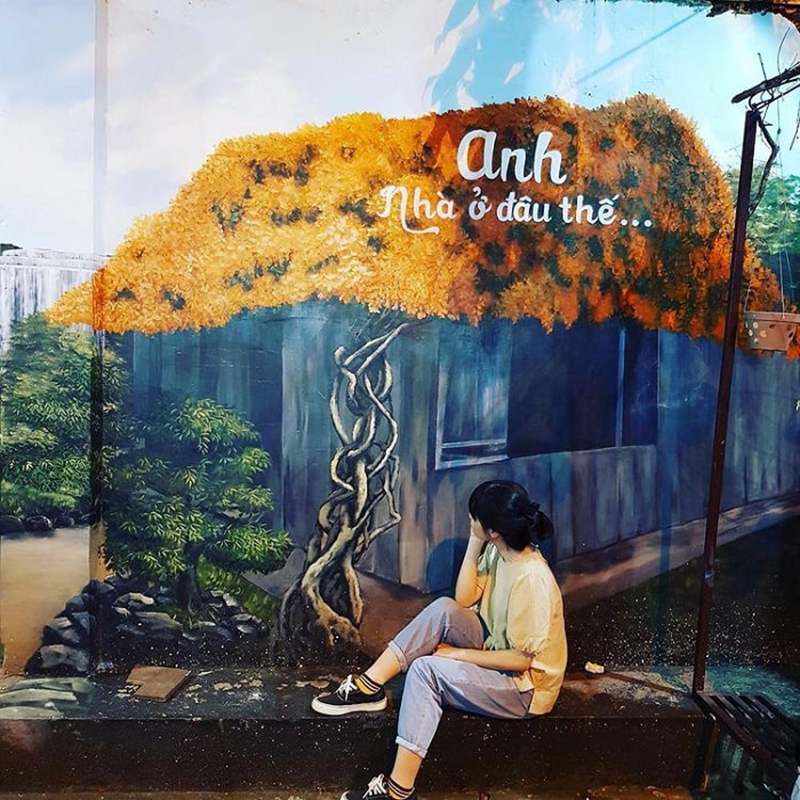 Người ngồi trước bức tranh tường mô tả ngôi nhà với cây leo và dòng chữ "Anh nhà ở đâu thế...".