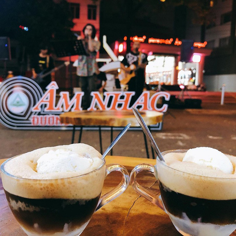 Hai ly cà phê kem trên bàn, phông nền là nhóm nhạc biểu diễn trên phố vào buổi tối.
