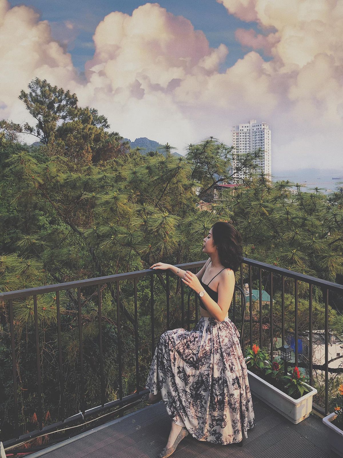 Người phụ nữ ngồi tr&ecirc;n ban c&ocirc;ng, nh&igrave;n ra quang cảnh c&acirc;y cối v&agrave; t&ograve;a nh&agrave; cao tầng dưới bầu trời nhiều m&acirc;y.