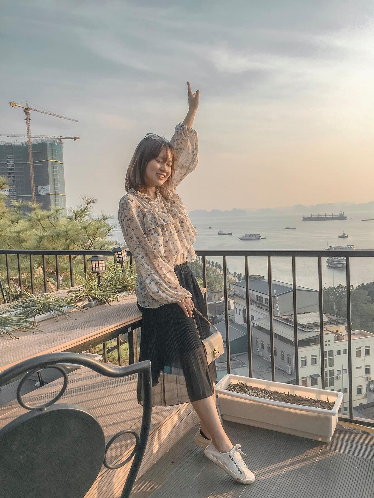 Người phụ nữ đứng tr&ecirc;n ban c&ocirc;ng, giơ tay l&ecirc;n, ph&iacute;a sau l&agrave; cảnh biển v&agrave; c&aacute;c t&ograve;a nh&agrave; dưới &aacute;nh ho&agrave;ng h&ocirc;n.