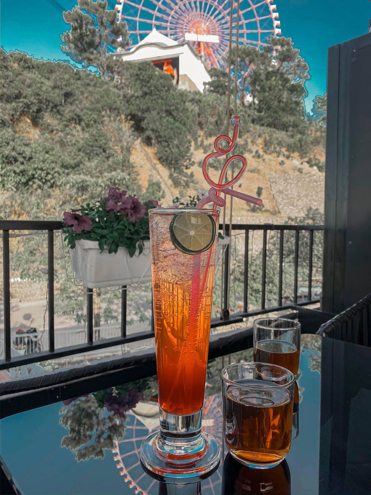 Ly nước &eacute;p cam v&agrave; hai ly tr&agrave; tr&ecirc;n b&agrave;n, ph&iacute;a sau l&agrave; cảnh quan đu quay v&agrave; c&acirc;y xanh.