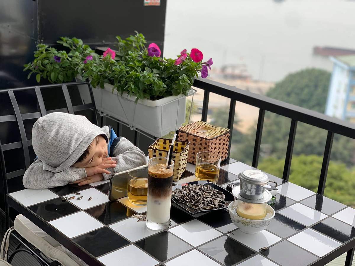 Một em b&eacute; đang ngủ gục tr&ecirc;n b&agrave;n ngo&agrave;i trời, b&ecirc;n cạnh l&agrave; đồ uống v&agrave; hoa trang tr&iacute;.