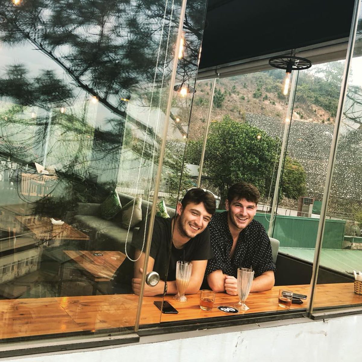 Hai người ngồi trong qu&aacute;n c&agrave; ph&ecirc;, mỉm cười nh&igrave;n ra ngo&agrave;i cửa sổ k&iacute;nh, với c&acirc;y cối v&agrave; đồi n&uacute;i ph&iacute;a sau.