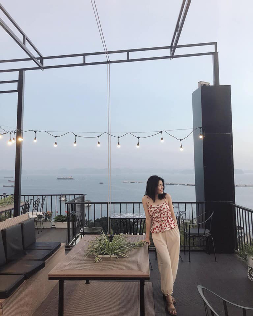 Một người đứng tr&ecirc;n s&acirc;n thượng, ph&iacute;a sau l&agrave; cảnh biển v&agrave; bầu trời, b&agrave;n ghế xung quanh v&agrave; d&acirc;y đ&egrave;n trang tr&iacute;.