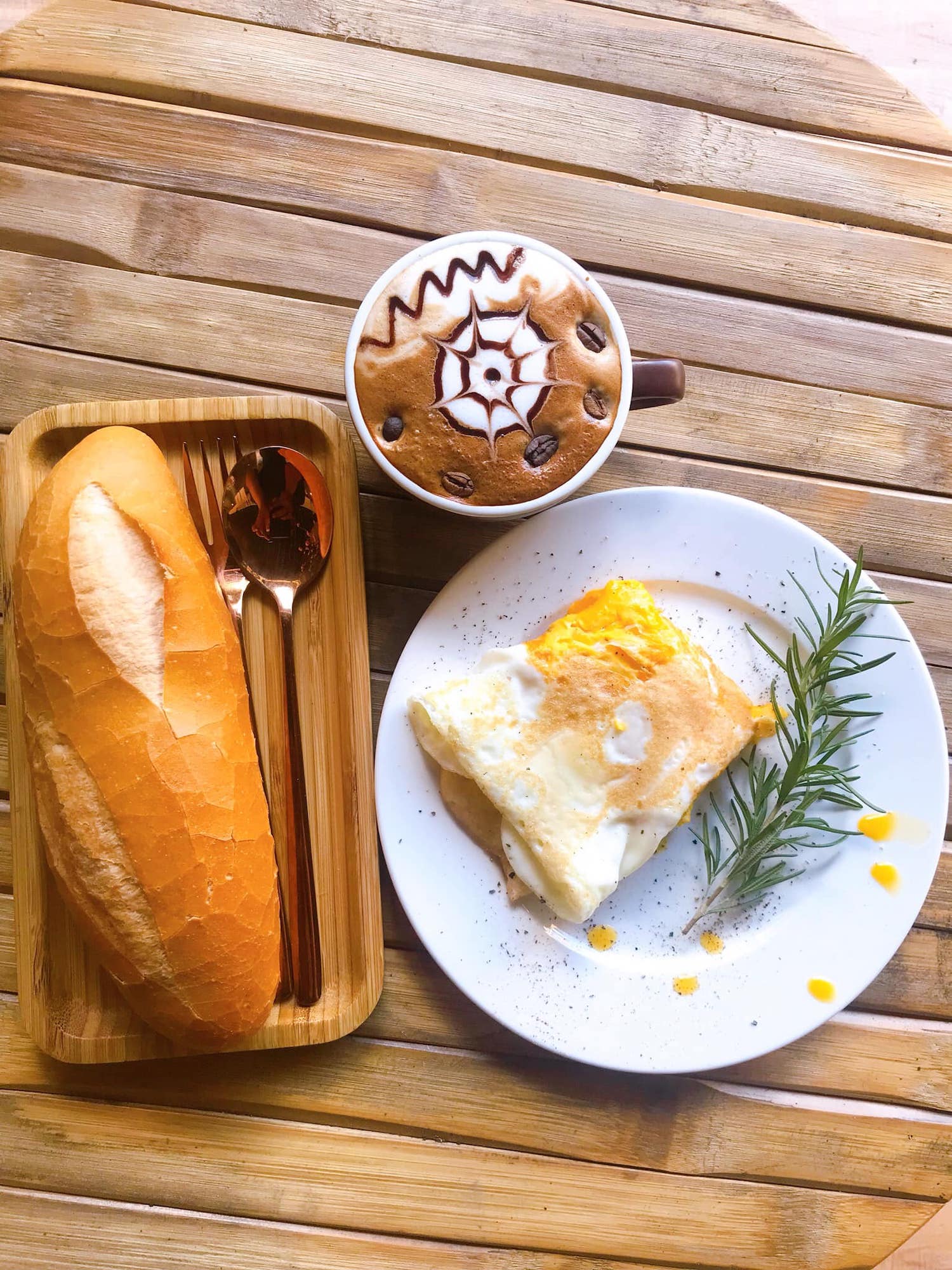 Bàn gỗ với ly cà phê nghệ thuật, bánh mì và đĩa trứng ốp la trang trí lá hương thảo.