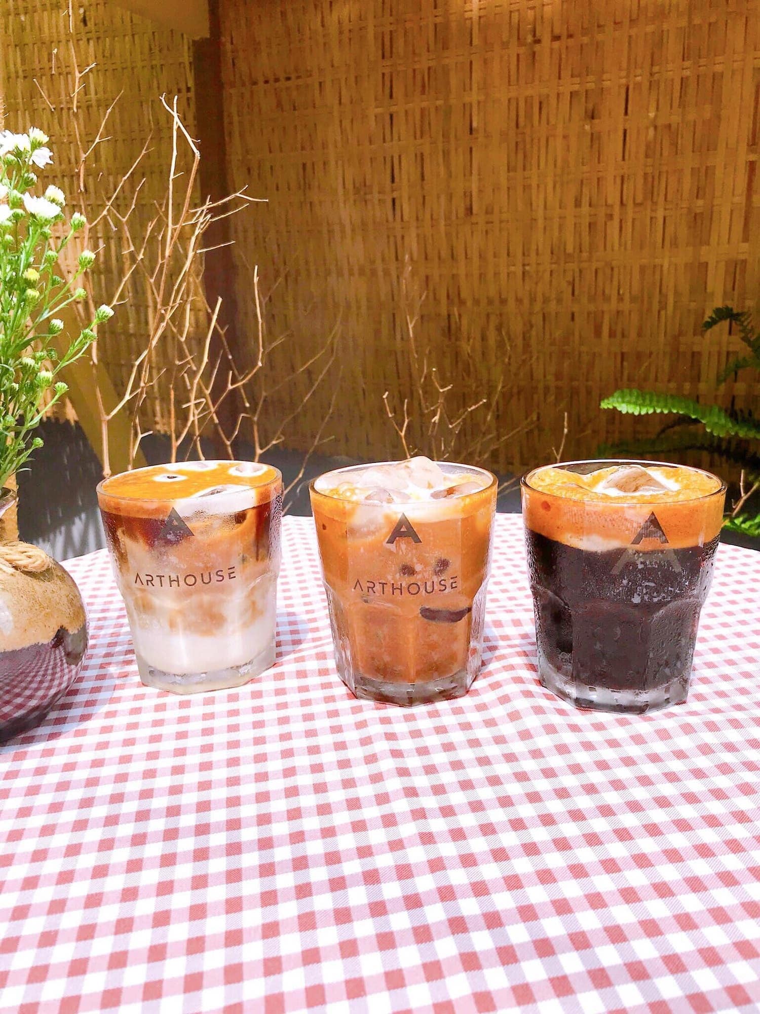 Ba ly cà phê đá đặt trên bàn trải khăn caro, nền tre, bên cạnh là bình hoa nhỏ.