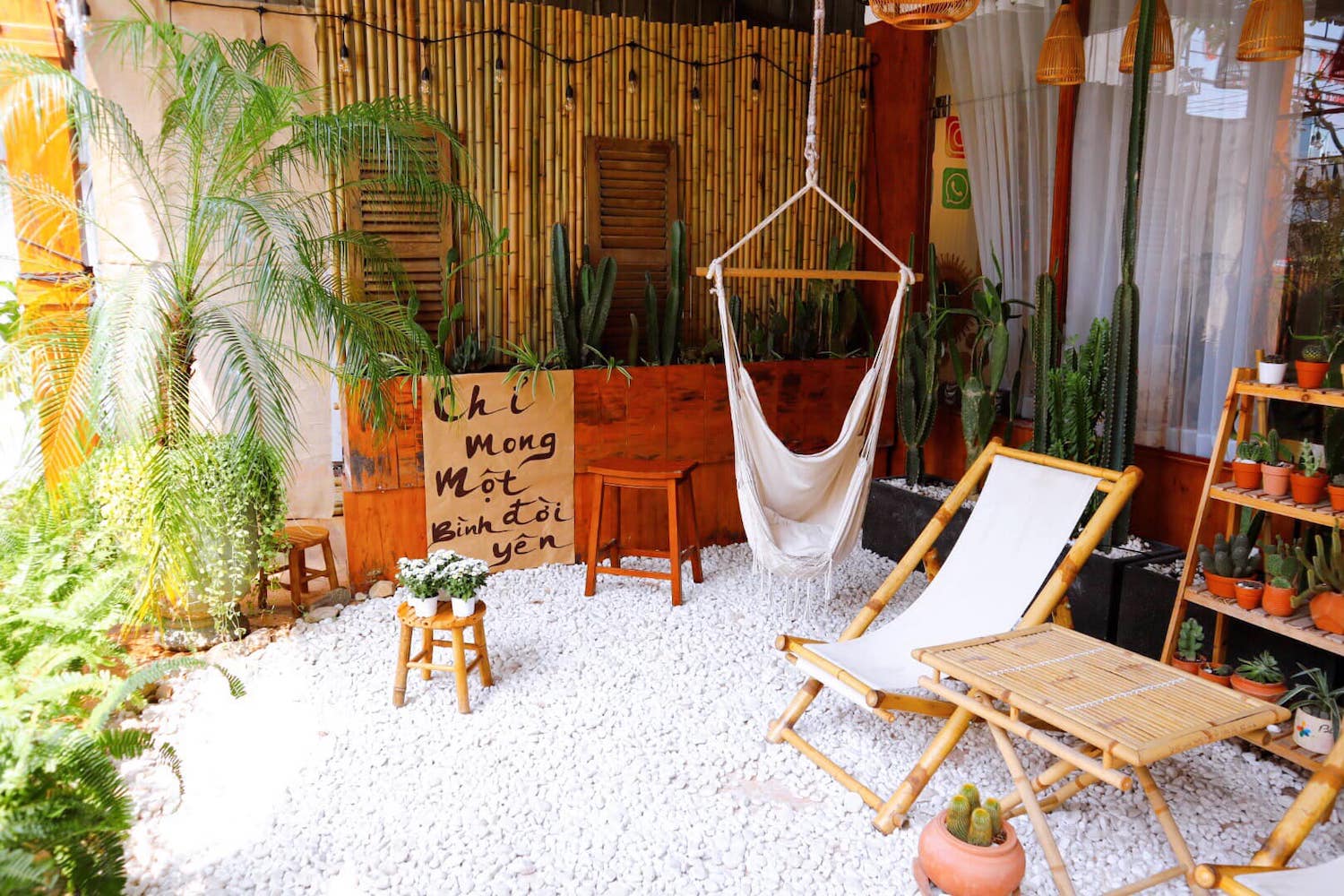Khu vườn nhỏ với ghế tre, võng, cây cảnh và bảng gỗ trên nền sỏi trắng, tạo không gian thư giãn.