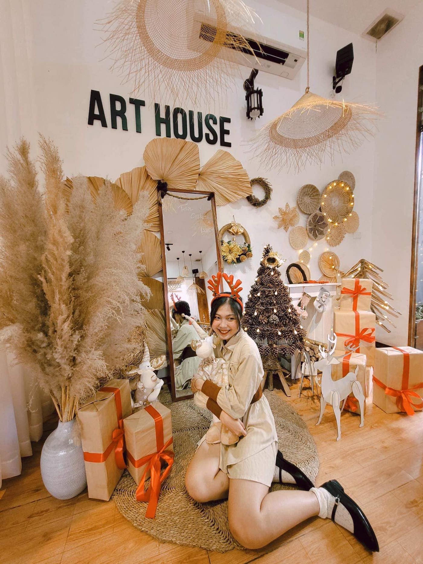 Người ngồi cạnh cây thông Noel và quà, trong không gian trang trí với đèn và phụ kiện nghệ thuật.